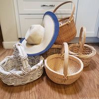 Five woven baskets and one woven straw hat displayed on hardwood floor showing shapes, sizes, and details of each item.