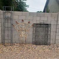 Photo showing three wrought iron garden pieces leaning against a cinder block wall on gravel ground: a narrow tall trellis, a rusted fan-shaped piece with leaf accents, and a rectangular fence section with decorative scrollwork.