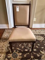 Front view of the wood chair showing beige upholstered seat and backrest, placed on patterned rug against wall.