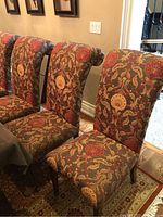 Photo showing three of the six armless upholstered dining chairs with rolled backs, lined up by a wall. Fabric has a floral and vine pattern in red, gold, beige, and gray tones.