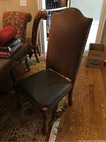 Front and side view of one brown faux leather chair with wood legs and nail head trim, placed indoors next to other patterned fabric chairs and hardwood floor.
