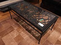 Front side angle view of the metal coffee table with glass top showing the decorative wrought iron pattern underneath and curved legs.