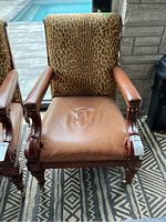 Front view of chair showing leather seat, leopard print fabric upholstered backrest, carved wood armrests and legs with brass nailhead trim.