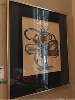 Framed botanical print showing artistic black ribbon-like design with botanical details, cream background and black mat inside gold-toned frame.