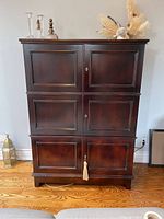 Front view of dark wood media cabinet showing three stacked sections with paneled doors and decorative key tassel on lower door. Cabinet stands on hardwood floor near wall with some decorative items on top.