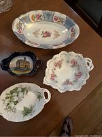 Four assorted plates shown on a wooden table including an elongated floral plate, a blue dish with building illustration, a scalloped floral plate, and a white floral plate with handles.