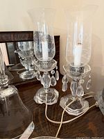 Image of two delicate crystal and glass electric lamps with dangling crystal prisms and glass hurricane shades, sitting on a wooden surface in front of a mirror.