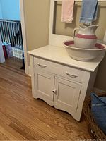 Front angle view of the white wooden wash stand showing the drawer, cupboard, and towel bar with two towels hanging and a pitcher and basin placed on top (not included in sale).