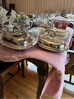 Two silver plate casseroles with lids shown on a table with other dining items, one rectangular and one oval shape, both with detailed handles and decorative edges.
