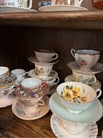 Assorted vintage tea cups and saucers stacked and displayed on wooden shelves with varied floral and colorful patterns visible.