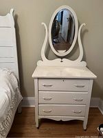 Photo of a small white painted dresser with three drawers and an attached oval mirror. Dresser has metal handles, curved wooden frame holding the mirror, and a slightly curved base raised on short legs.
