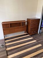 Full-size wooden bed frame with headboard that has open cubby storage, wood slats for mattress support, next to a matching medium-brown wooden dresser with six drawers and carved design on the lower front.