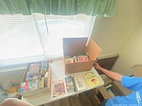 Books arranged in cardboard boxes and on a table near window, including cookbooks, fiction, and non-fiction books visible.