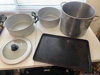 Cookware set displayed on white table showing large stockpot, steamer insert, smaller pot, glass lid, and baking sheet.
