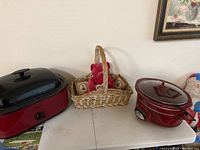 Two red Crockpots, one with black lid and one with clear lid sitting on a white table with a basket between them.