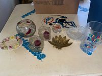 Image showing nine glassware items including various candle holders, a decorative glass ball, a brass lotus-shaped holder, a decorative floral plate, and a tall glass cup with colorful text.