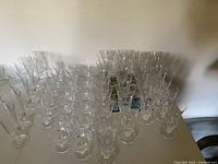 Photo showing an assortment of clear crystal glassware, including multiple wine glasses and cups, arranged on a flat surface against a wall.