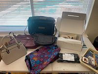 Wide shot of all handbags, fabric bag, boxed shoes, cutlery tray, and sandals displayed on a table in front of a window.