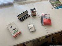 Overview of the lot including Bluetooth headphones, boxed pens, collectible matchbox, playing cards, and cigarette papers on a table