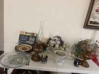 Collection of vintage interior decorations including oil lamps, covering plates, floral arrangements, and glassware on white table with painting hung on the wall