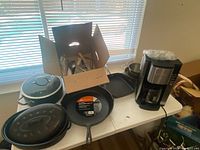 Photo showing coffee maker, pots, pans, cutlery box and utensils on table near window.
