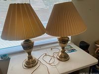 Pair of vintage brass table lamps with pleated beige shades on white table in front of window.