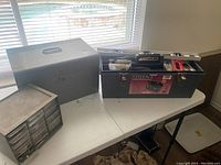 Wide shot showing two metal toolboxes on a white table near a window. One toolbox is open showing various hand tools and hardware inside.