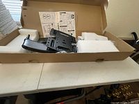 Open box displaying the black TV wall mount with mounting bracket, mounting hardware, white foam packaging inserts, and instruction manual.