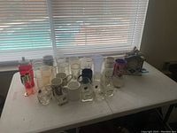 Table with a variety of vintage glassware including clear drinking glasses, white and colored mugs, a pink translucent water bottle, and a decorative house-shaped clock.