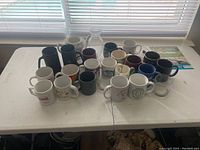 Table with an assortment of 24 vintage glassware and ceramic mugs, including black, white, maroon colored ceramic mugs and clear glass mugs.