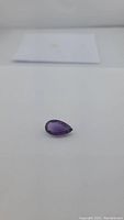 Close-up image of a deep purple pear-shaped amethyst gemstone placed on a plain surface.