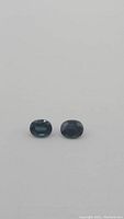 Pair of natural teal blue-green oval sapphires placed side by side on white background.