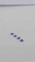 Four pear-shaped blue purple tanzanite gemstones arranged in a line on a plain background, showing size and color.