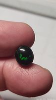 Close-up of the natural jet black oval Ethiopian opal cabochon held between fingers, showing vivid green color flashes against a deep black background.