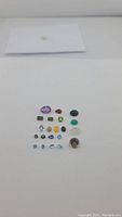 Front view of a parcel of 22 assorted natural gemstones of various cuts and colors arranged on a white surface.