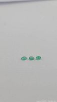 Photo of three oval natural Zambian emerald gemstones displayed in a row on a plain background.
