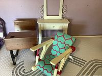 Photo shows four pieces of American Girl doll furniture arranged indoors against a wall. Includes a wooden school desk, cream-colored side table with drawer, fabric cushioned reclining chair, and a doll bed frame.