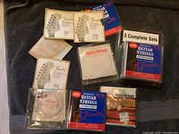 Photo showing assorted new guitar strings packages laid out on a dark fabric background including Fender, Darco, and other professional electric guitar strings.