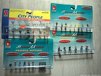 Four packages of HO scale hand-painted model figures arranged on floor. Top left package titled City People with 8 figures visible. Top right two packages titled Scene Master Townspeople and Railroad Workers, each with 8 figures. Bottom package is Scene Master People Sitting with 8 figures including seated figures and a small bench.