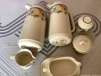 Top view of two tall Mikasa coffee pots with peach and leaf design, two small lidded jars and two small pitchers with blueberry and peach patterns.