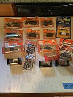 Photo shows 14 boxed and packaged collectible miniature vehicles and tanks, mostly Minitanks and Matchbox models, arranged on a white surface.