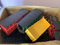 Train cars and tracks arranged loosely inside a cardboard box with some overlapping cars and visible wires.