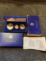 Three commemorative coins in blue velvet case with lid open, outer sleeve, and certificate sheet visible