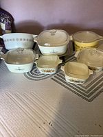 View of entire lot showing seven casseroles of different sizes and patterns including Corningware Blue Cornflower, floral vegetable motif, and Pyrex yellow starburst bowl.
