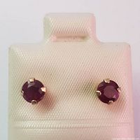 Pair of 10K yellow gold stud earrings with round ruby gemstones in prong settings displayed on a white textured holder.