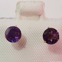 Front view of both round, faceted amethyst gemstones in yellow gold four-prong settings shown on white textured display.