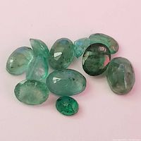 Photo of 10 faceted emerald gemstones laid out on a white background showing color and cut variations.