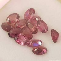 Close-up photo showing fourteen loose faceted garnet gemstones with oval and pear shapes in deep pink to reddish color.