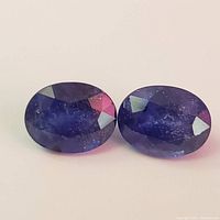 Close-up front view of two oval cut blue sapphires placed side by side on a white background showing facets and color.
