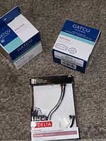 Three boxed chrome robe hooks (two Gatco, one Delta) on carpet.
